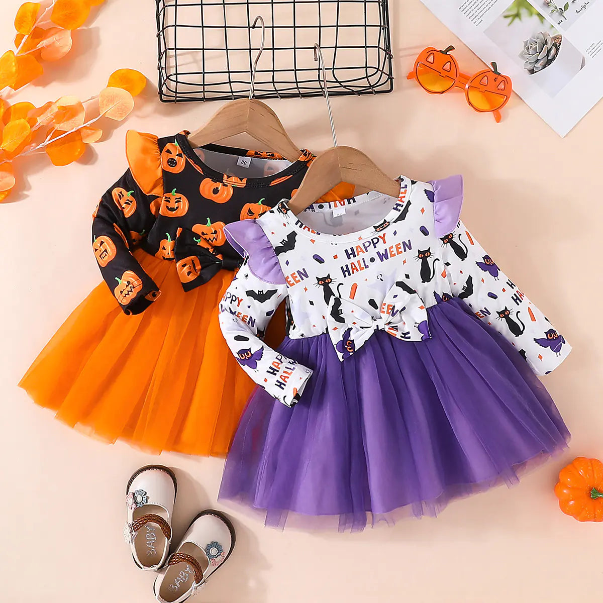 Two Halloween-themed children's dresses on hangers with accessories on a pink background.